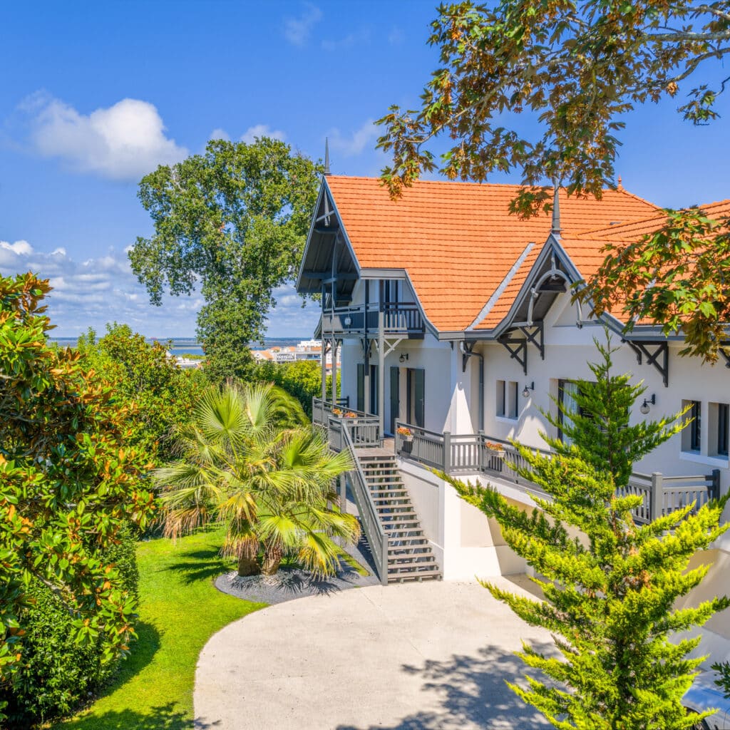 Villa de Prestige à Arcachon avec Vue Mer et Grand Jardin Villa de luxe à Arcachon avec toit rouge, balcons gris et jardin luxuriant avec palmier sous un ciel bleu.